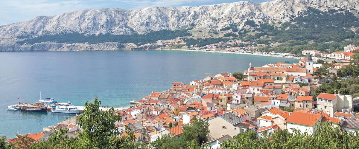 A coastal town with red-roofed buildings, boats docked at the harbor, and a mountain range in the background.