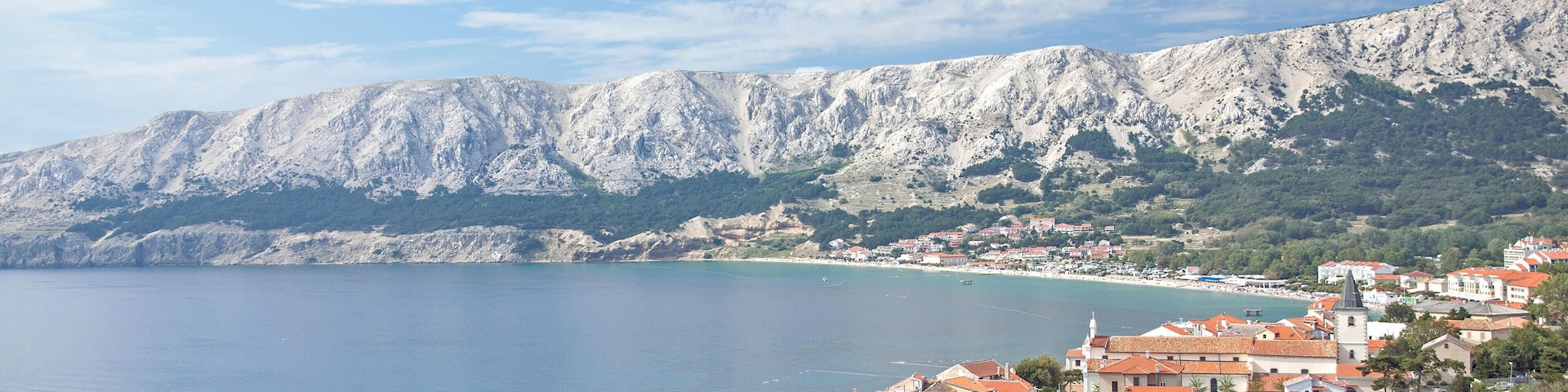A coastal town with red-roofed buildings, boats docked at the harbor, and a mountain range in the background.