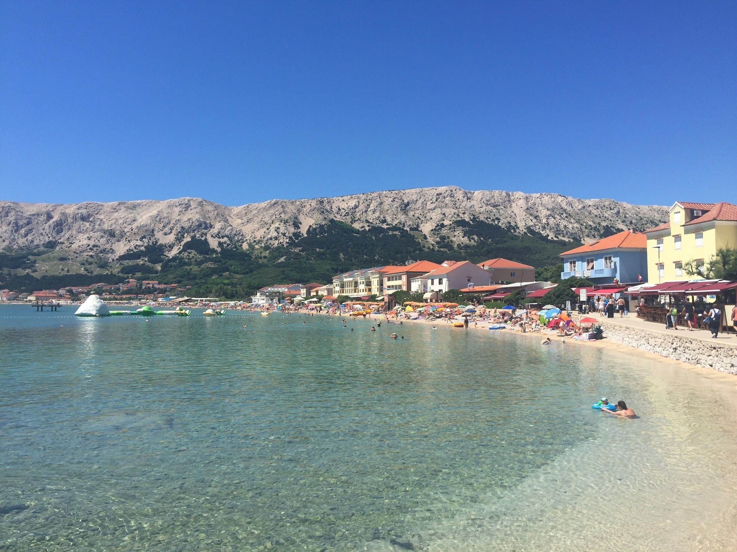 A clear coastal beach with buildings and mountains in the background.