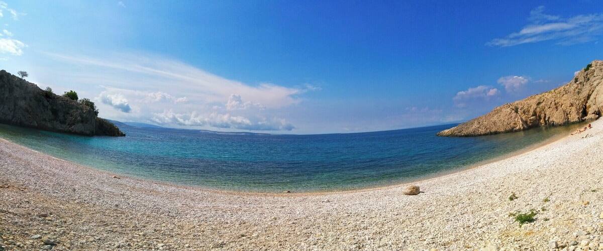 A pebble beach with clear turquoise water, rocky cliffs, and a clear sky.