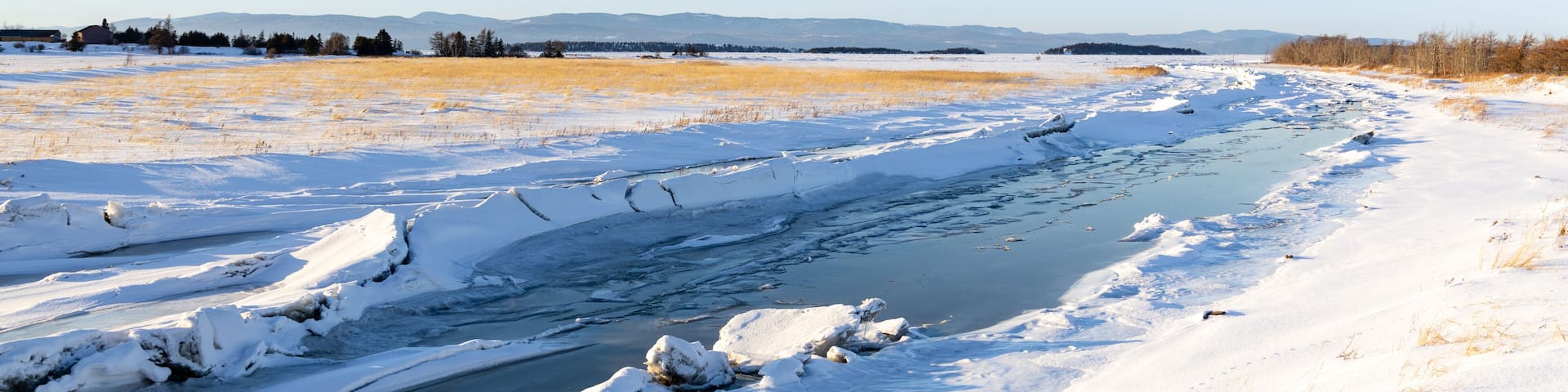 The partly frozen Kamouraska river flowing through snowy fields into the St. Lawrence River seen during a golden hour winter afternoon, Kamouraska, Quebec, Canada