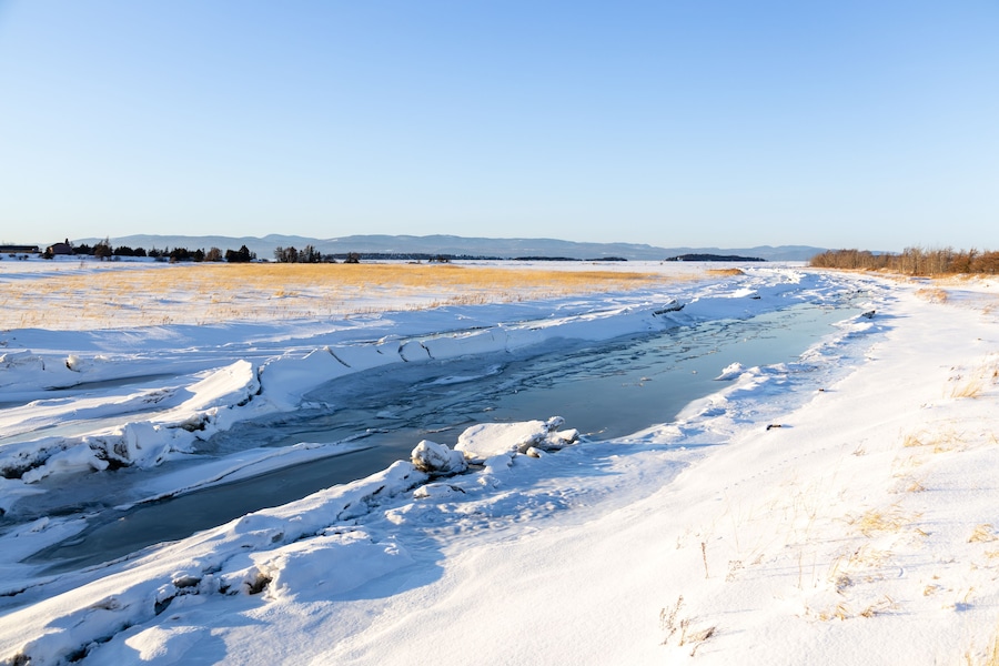 The partly frozen Kamouraska river flowing through snowy fields into the St. Lawrence River seen during a golden hour winter afternoon, Kamouraska, Quebec, Canada