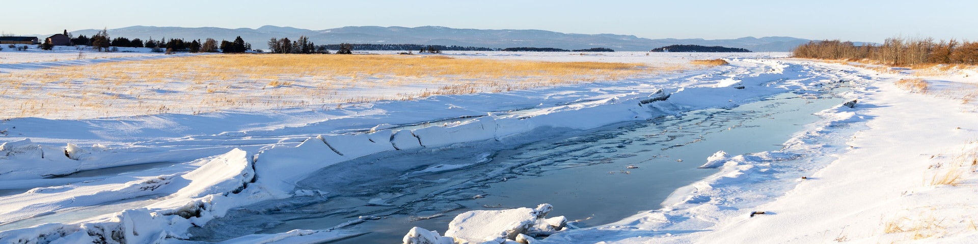 The partly frozen Kamouraska river flowing through snowy fields into the St. Lawrence River seen during a golden hour winter afternoon, Kamouraska, Quebec, Canada