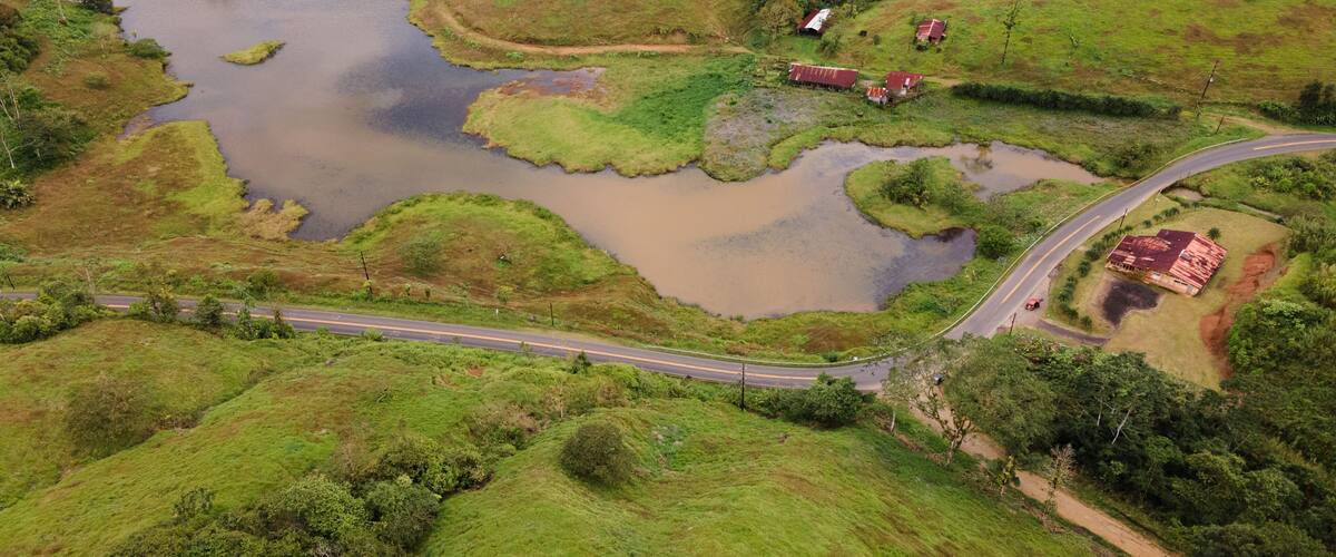 An aerial view of a rural landscape with a winding road, a small body of water, and a few scattered buildings.