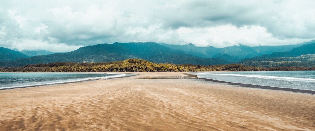A beach with a sandy shore, calm water, and a backdrop of mountains and dense forest.