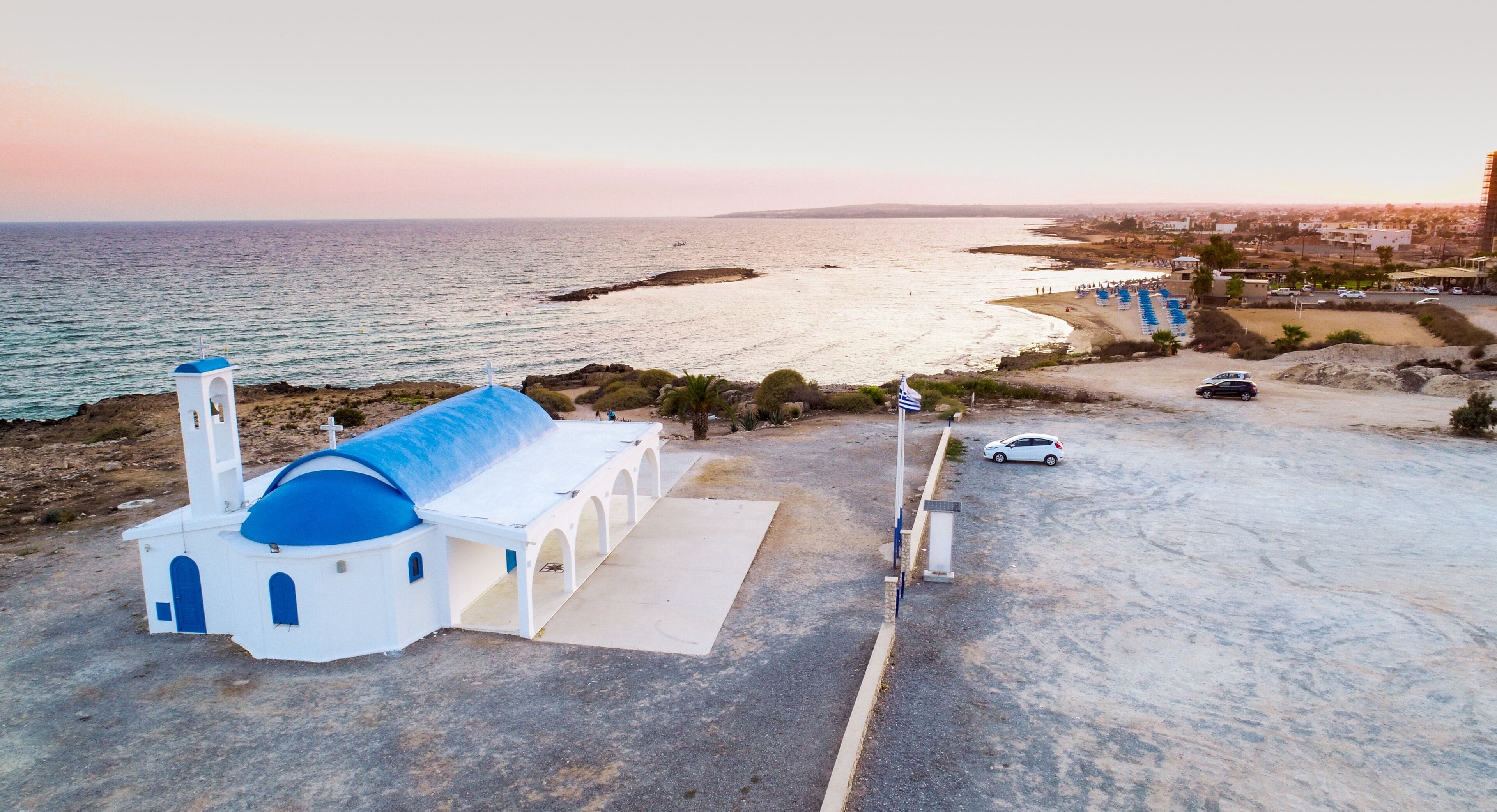 Aerial bird's eye view of coastline sunset, traditional landmark white and blue chapel at Agia Thekla beach, Ayia Napa, Famagusta, Cyprus from above.Tourist attraction golden sand bay, church, sunbeds