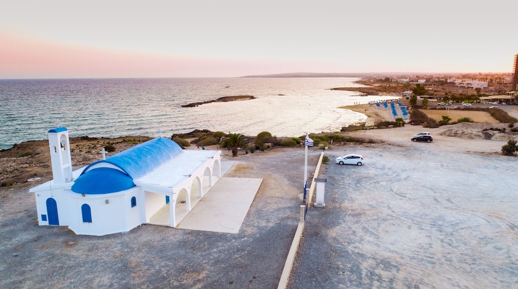 Aerial bird's eye view of coastline sunset, traditional landmark white and blue chapel at Agia Thekla beach, Ayia Napa, Famagusta, Cyprus from above.Tourist attraction golden sand bay, church, sunbeds