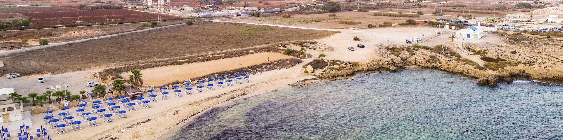 Aerial bird's eye view of coastline sunset, traditional landmark white and blue chapel at Agia Thekla beach, Ayia Napa, Famagusta, Cyprus from above.Tourist attraction golden sand bay, church, sunbeds