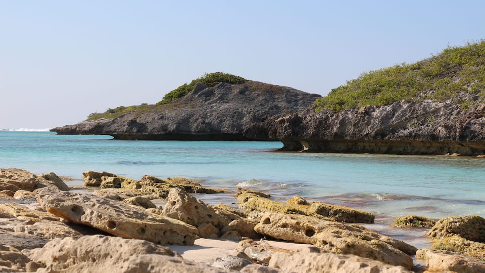 A rocky beach with clear turquoise water and a large rock formation with sparse vegetation.