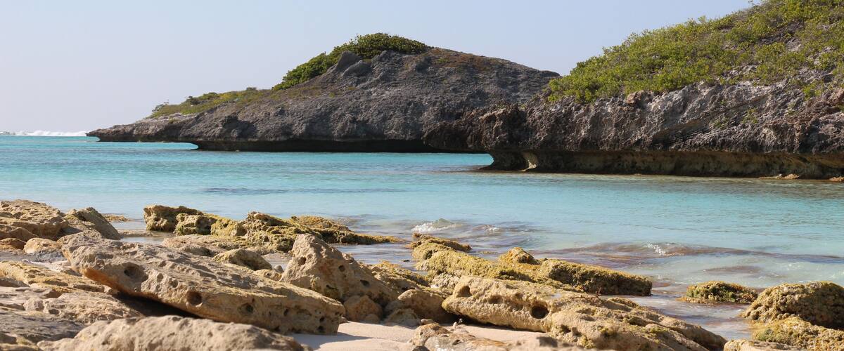 A rocky beach with clear turquoise water and a large rock formation with sparse vegetation.