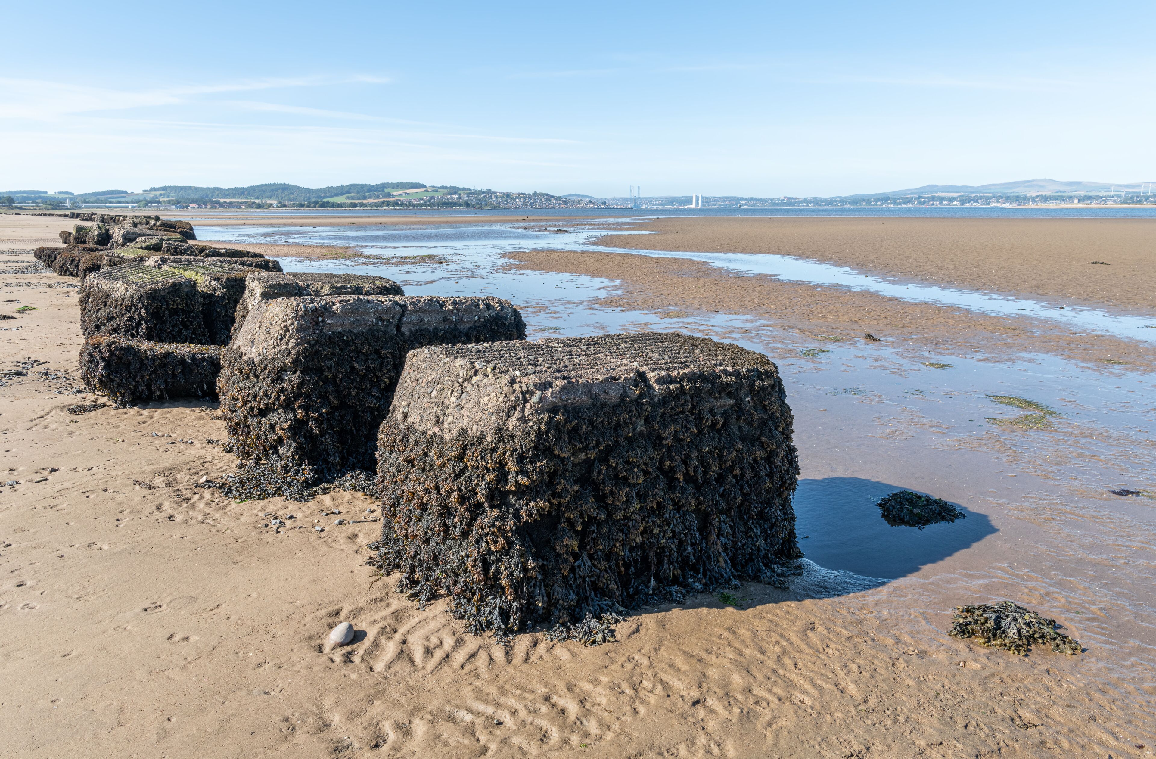 A sandy beach with large, cylindrical rocks and a distant view of a bridge and hills.