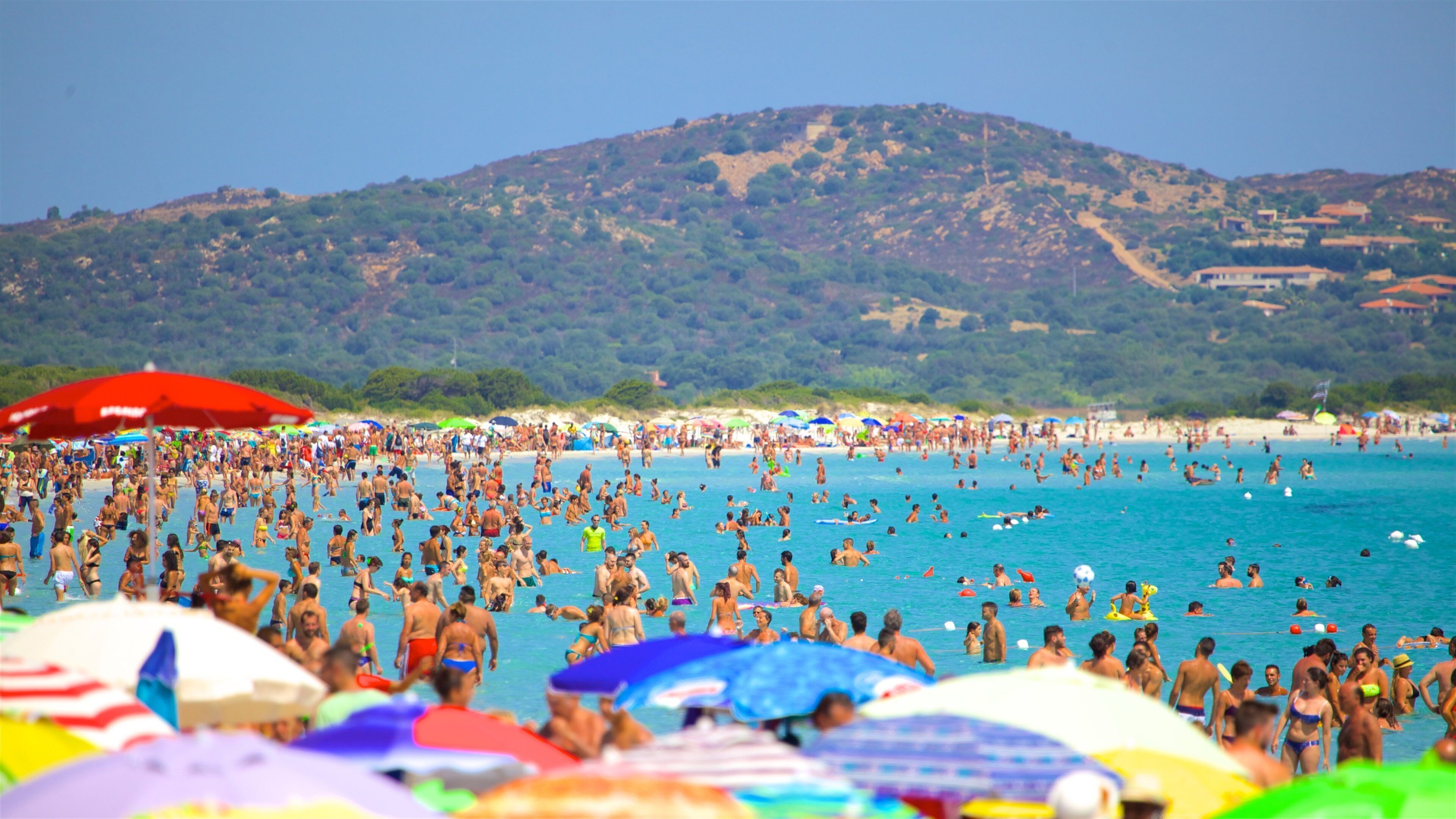 A crowded beach with people swimming and sunbathing, colorful umbrellas, and a mountainous backdrop.