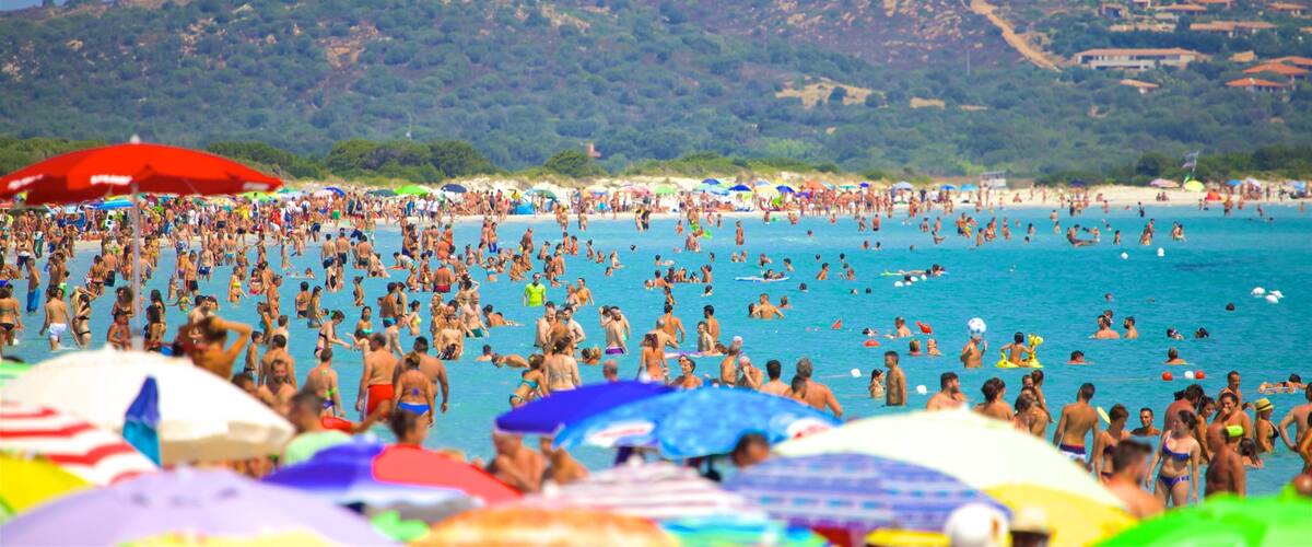 A crowded beach with people swimming and sunbathing, colorful umbrellas, and a mountainous backdrop.