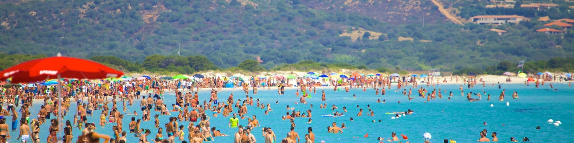 A crowded beach with people swimming and sunbathing, colorful umbrellas, and a mountainous backdrop.