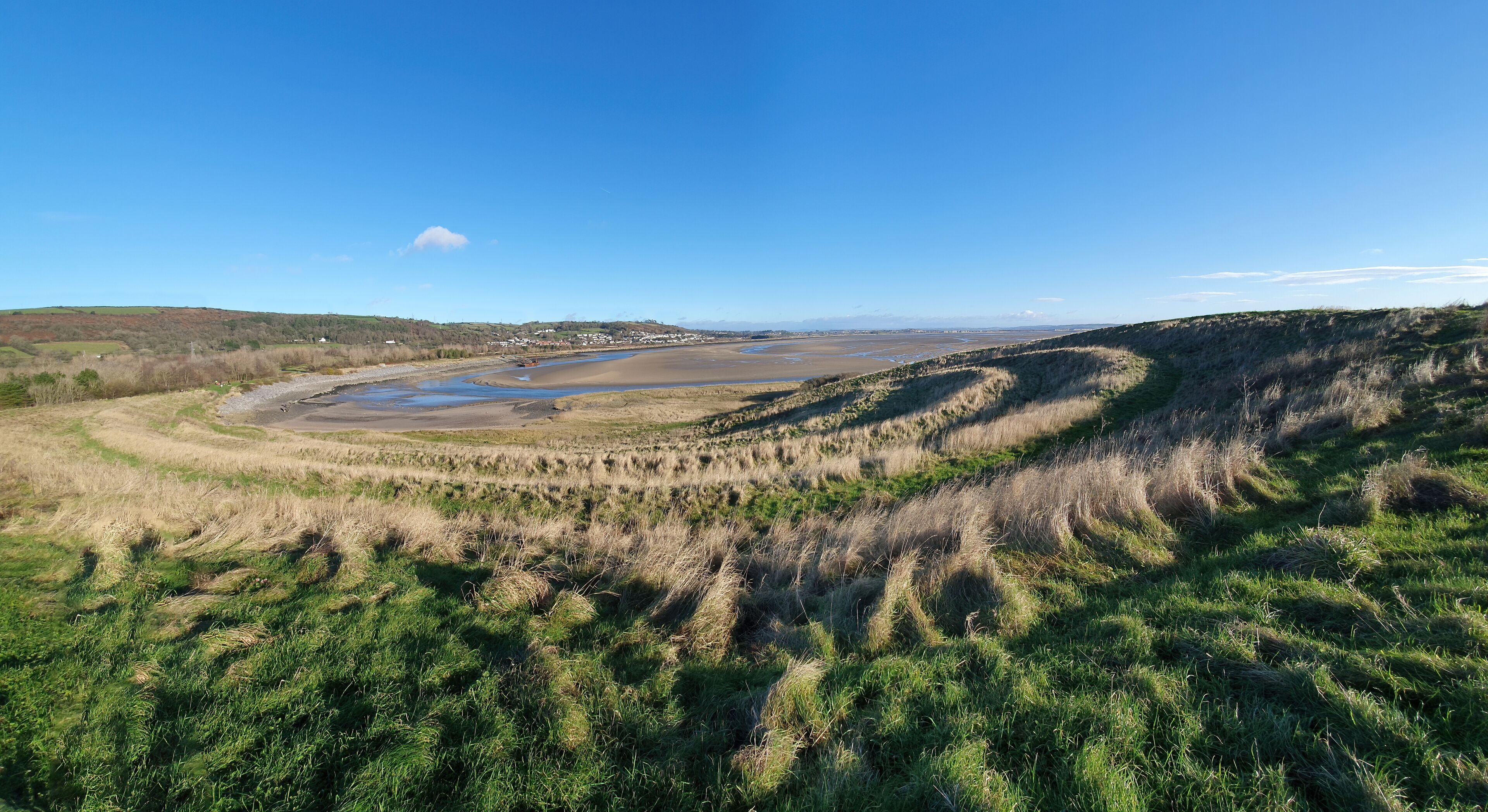 A coastal landscape with a river, grassy dunes, and distant hills.