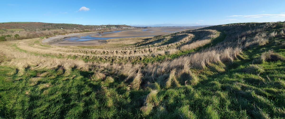 A coastal landscape with a river, grassy dunes, and distant hills.