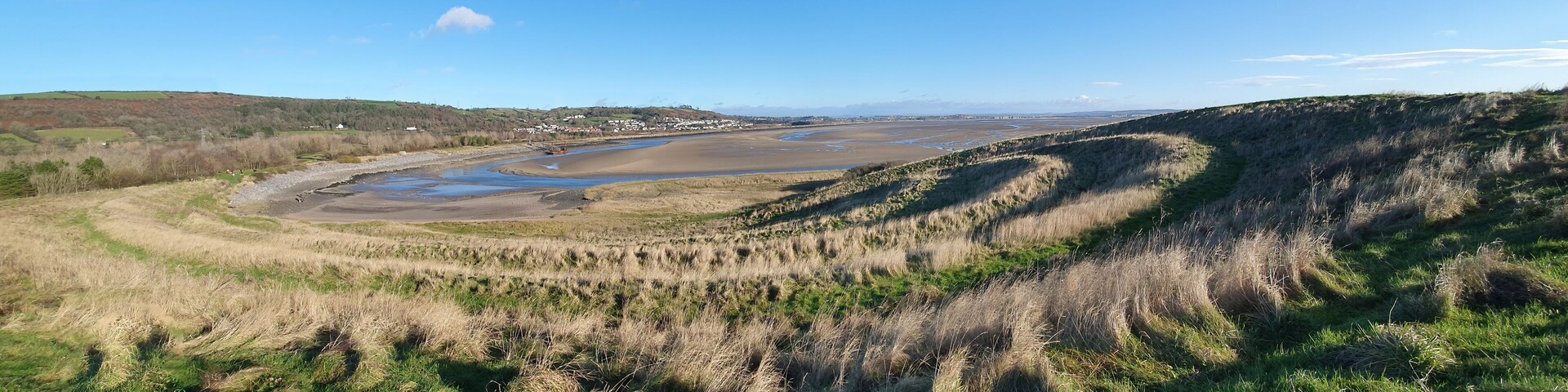 A coastal landscape with a river, grassy dunes, and distant hills.