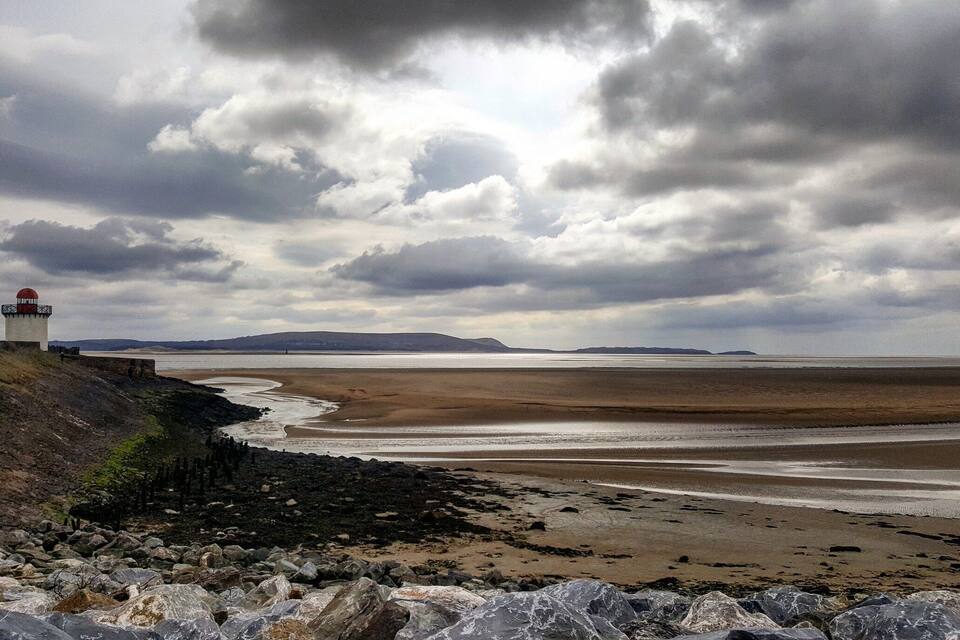 A coastal landscape with a lighthouse, rocky shore, and a wide beach under a cloudy sky.