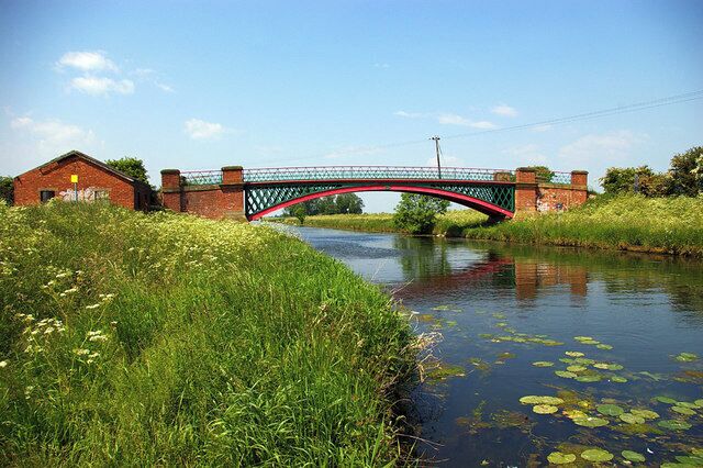 A red and green bridge over a canal with water lilies, surrounded by greenery and a brick building.