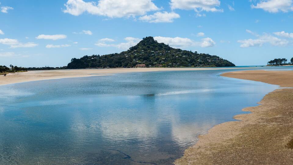 A beach with calm waters, a sandy shore, and a hill with buildings in the background.