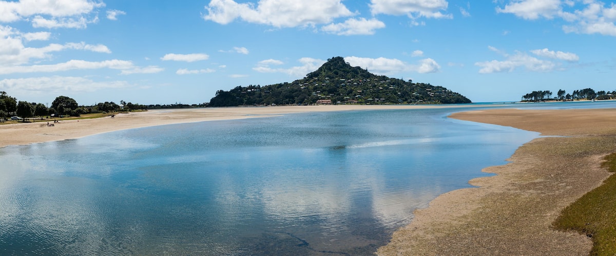 A beach with calm waters, a sandy shore, and a hill with buildings in the background.