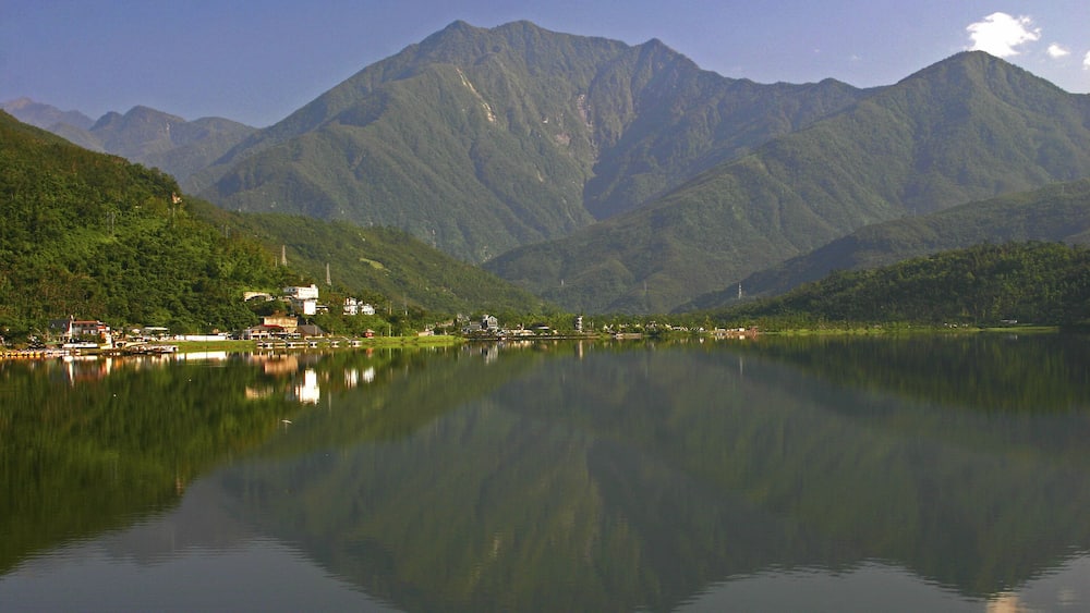 A calm lake with a mountain range in the background and a small settlement on the left.