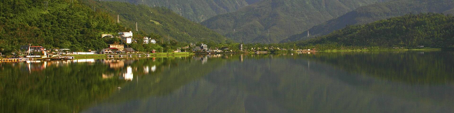 A calm lake with a mountain range in the background and a small settlement on the left.