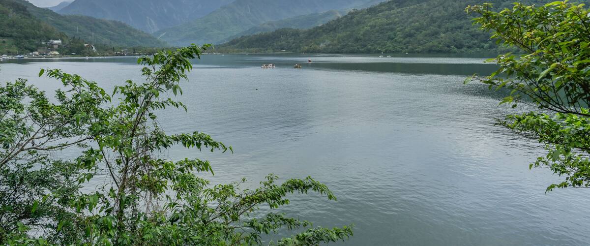 A calm lake surrounded by lush green hills and mountains under a cloudy sky.