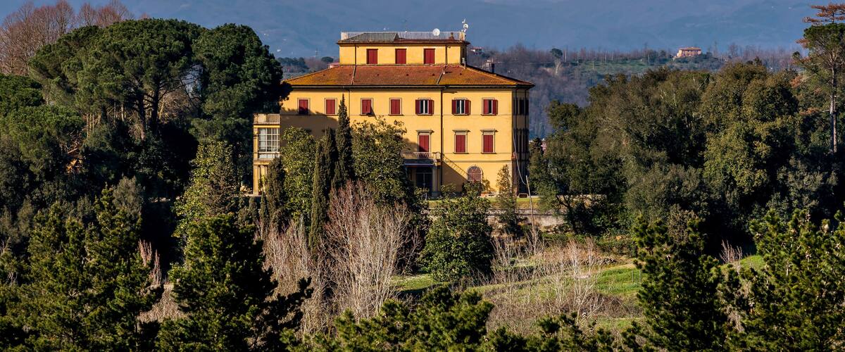 A yellow villa with red shutters surrounded by trees, with snow-capped mountains in the background.