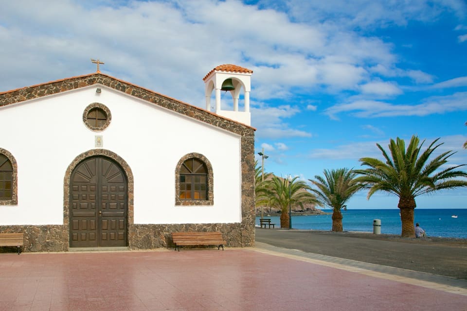 Uma igreja branca com um campanário, duas janelas em arco e uma grande porta de madeira. Há bancos em frente ao edifício, palmeiras e um corpo d'água ao fundo.