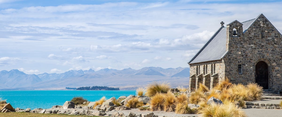 A stone church with a bell tower, surrounded by dry grass and mountains in the background.