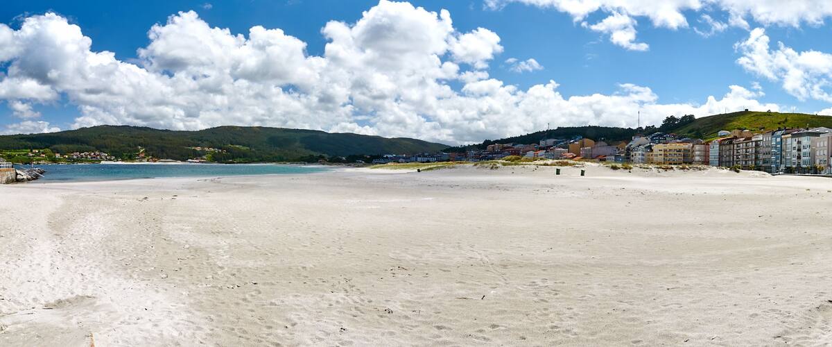 A wide sandy beach with a clear blue sky and scattered clouds. In the background, there are buildings along the coastline and a green hill with trees.