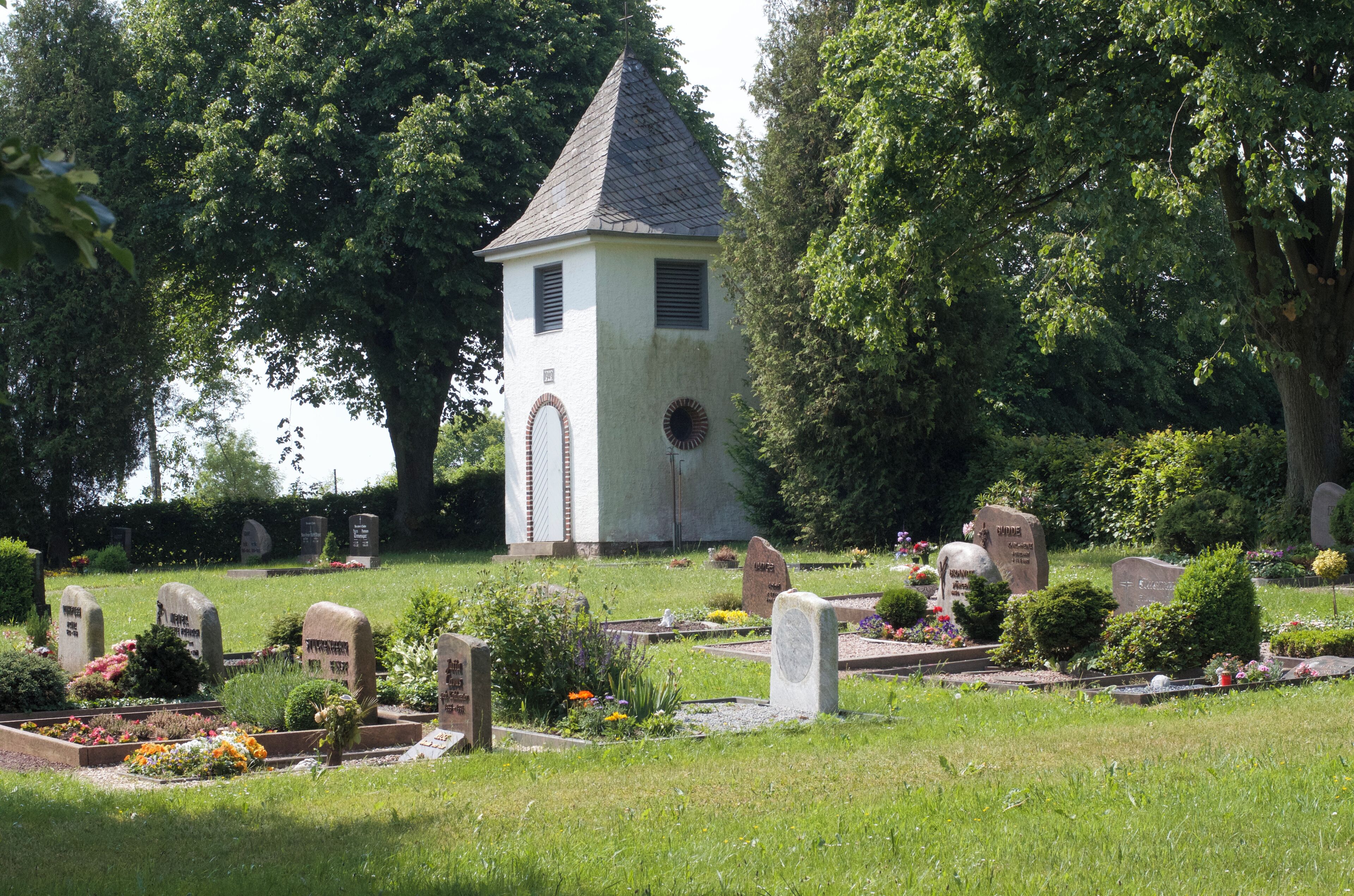 Blick über das Gräberfeld auf den Turm zu. Der Friedhof ist von einer Hecke gesäumt und hat zwei Eingänge zur Straße.