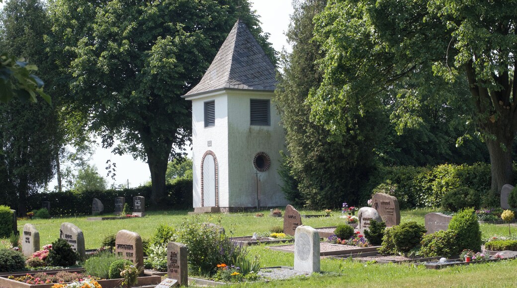 Blick über das Gräberfeld auf den Turm zu. Der Friedhof ist von einer Hecke gesäumt und hat zwei Eingänge zur Straße.