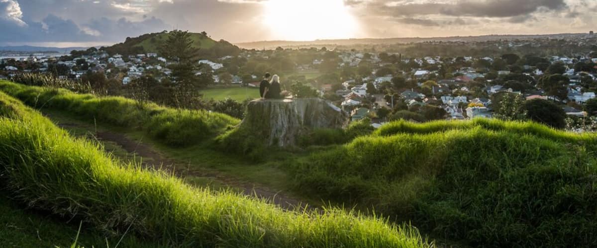 A grassy hill with a large rock, a town in the background, and a partly cloudy sky with the sun setting or rising.