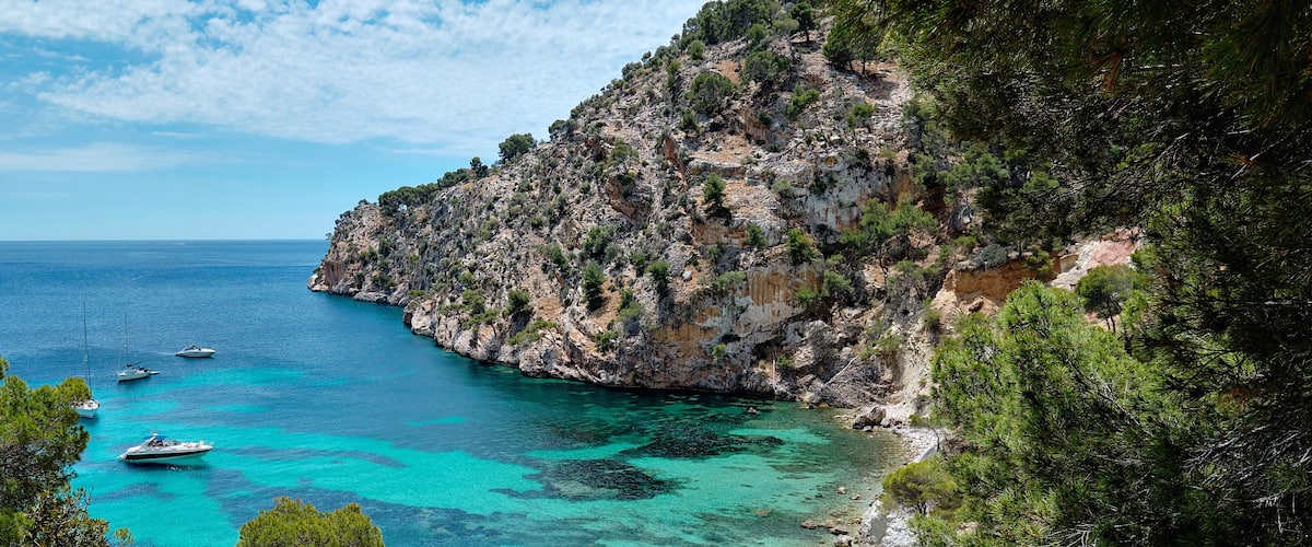 A coastal landscape with clear turquoise waters, rocky cliffs, and boats anchored near the shore.