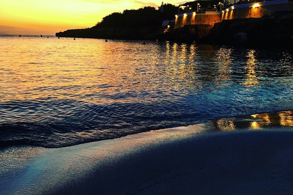 A sunset over a calm sea with a beach and a building with palm trees.