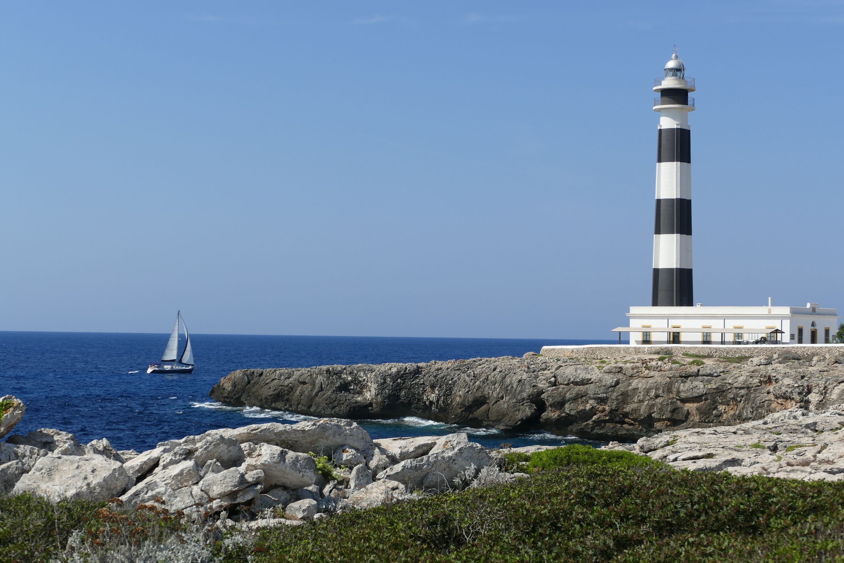 A lighthouse, rocky coastline, and a sailboat on the sea.