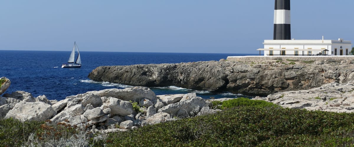 A lighthouse, rocky coastline, and a sailboat on the sea.