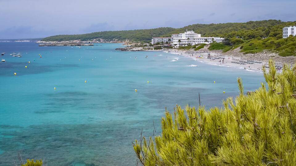 A coastal beach with clear turquoise waters, a sandy shore, and a building on a hillside.