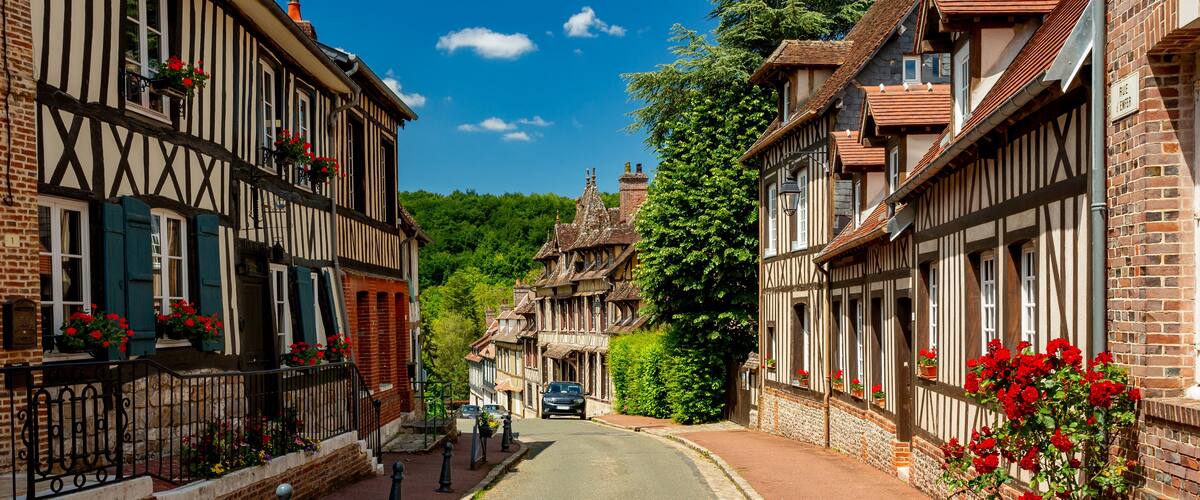 A narrow street lined with half-timbered houses, red flowers, and a car parked in the distance.