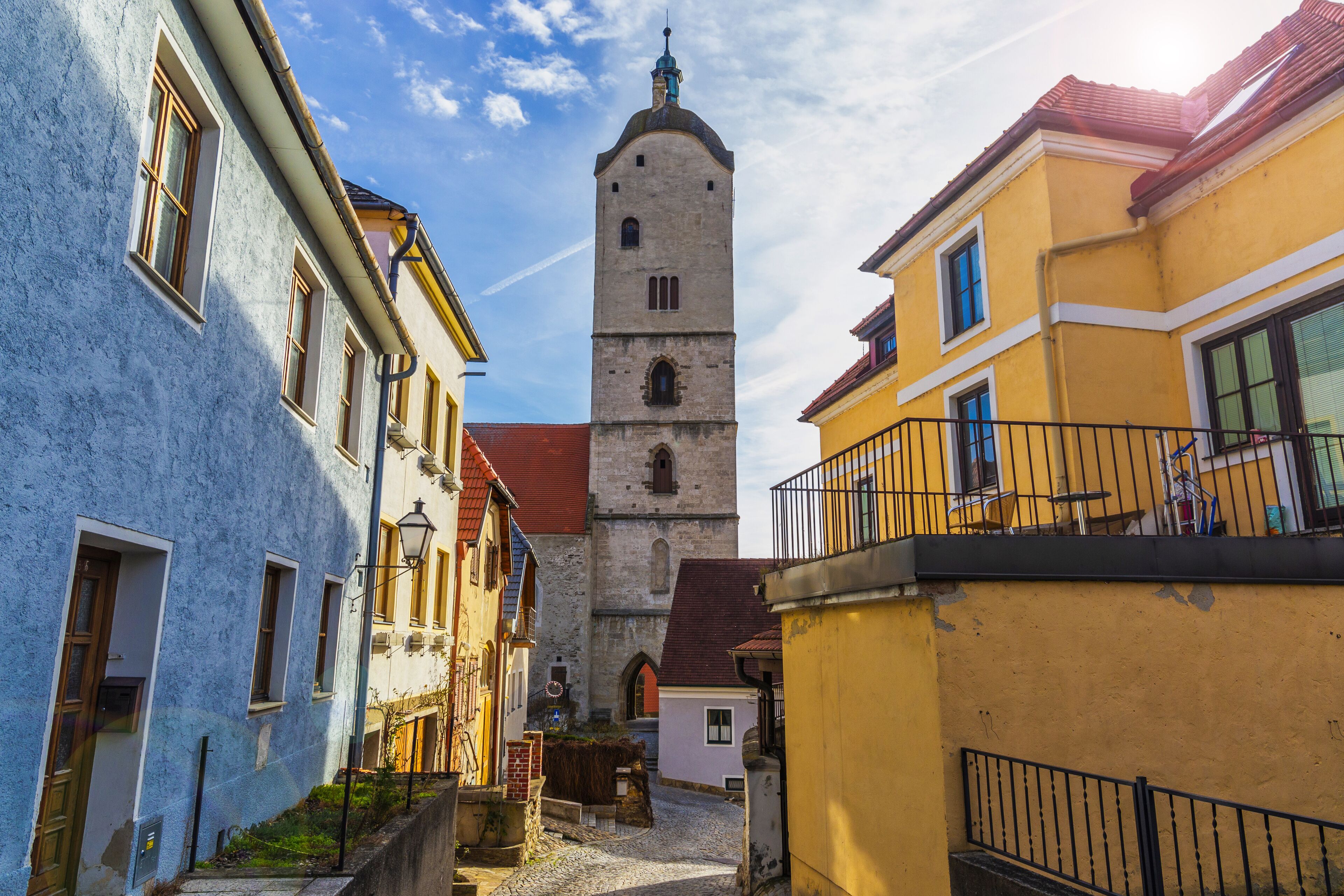 A narrow street with a historic tower, colorful buildings, and cobblestone pavement.