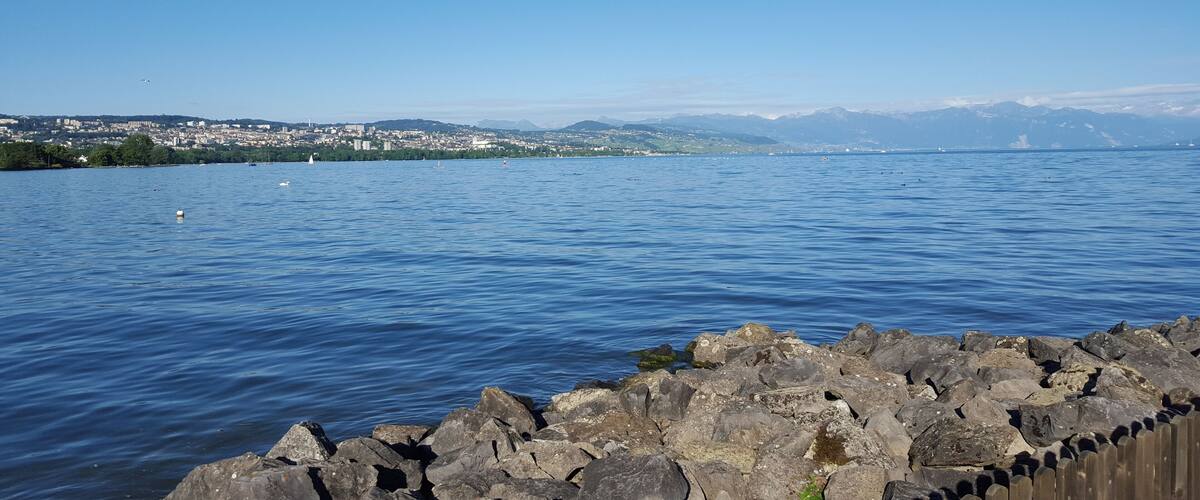 A calm lake with a rocky shoreline, a distant cityscape, and mountains in the background.