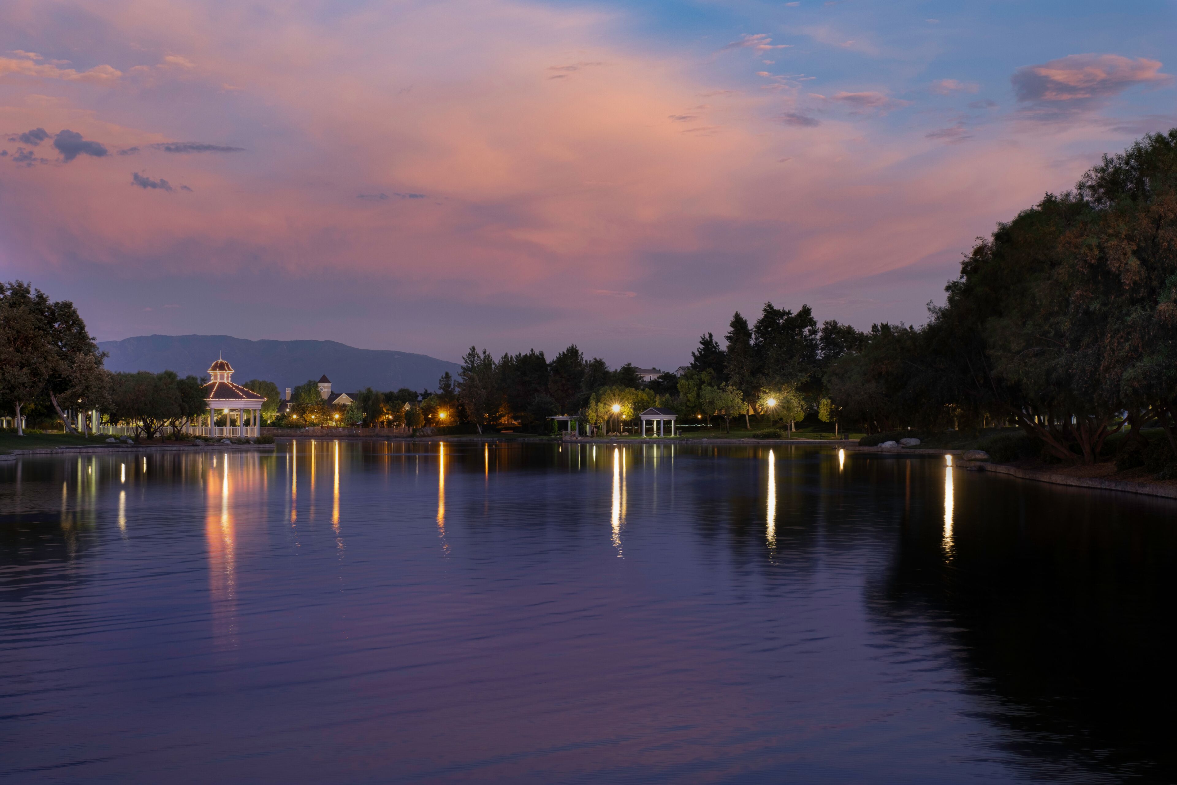 A calm lake at dusk with a gazebo and trees reflecting on the water.