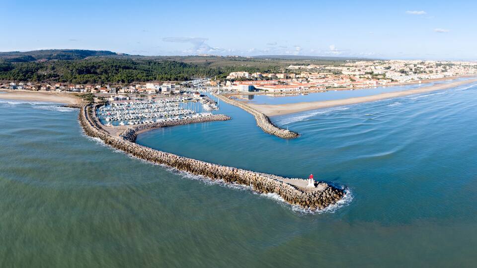 A coastal marina with boats, a lighthouse, and a beach with buildings in the background.