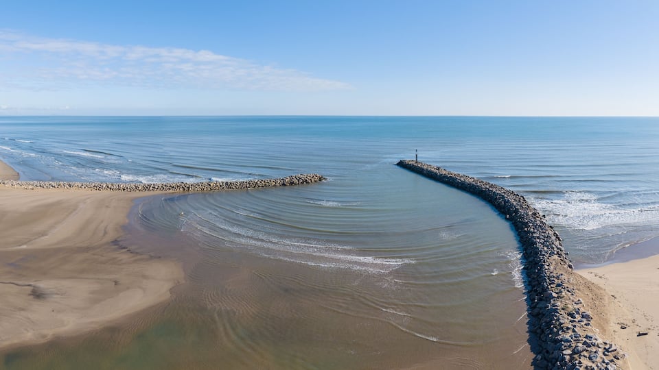 A coastal landscape with a curved breakwater extending into calm waters, a sandy beach, and a clear sky.