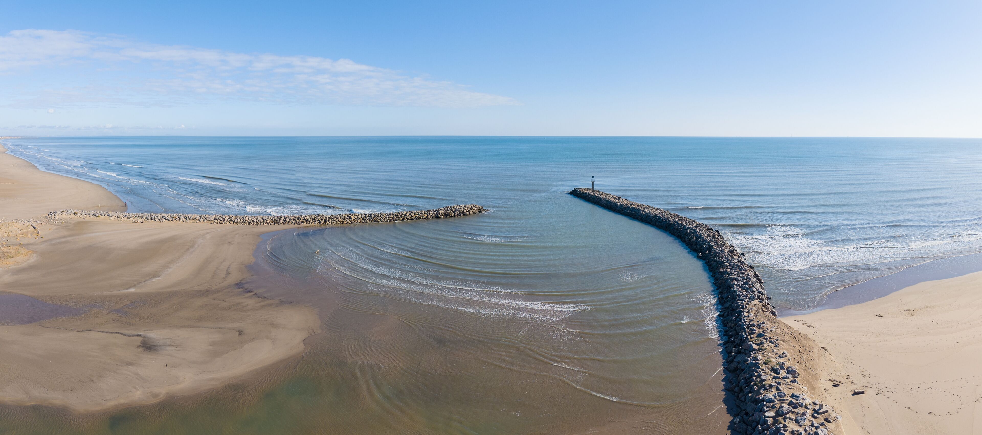 A coastal landscape with a curved breakwater extending into calm waters, a sandy beach, and a clear sky.