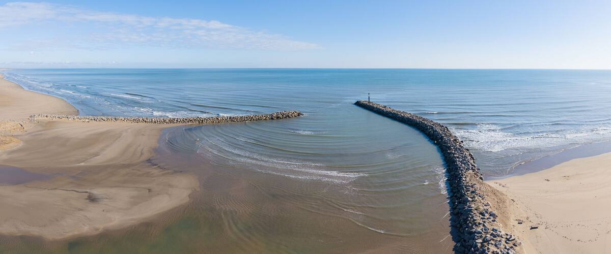 A coastal landscape with a curved breakwater extending into calm waters, a sandy beach, and a clear sky.
