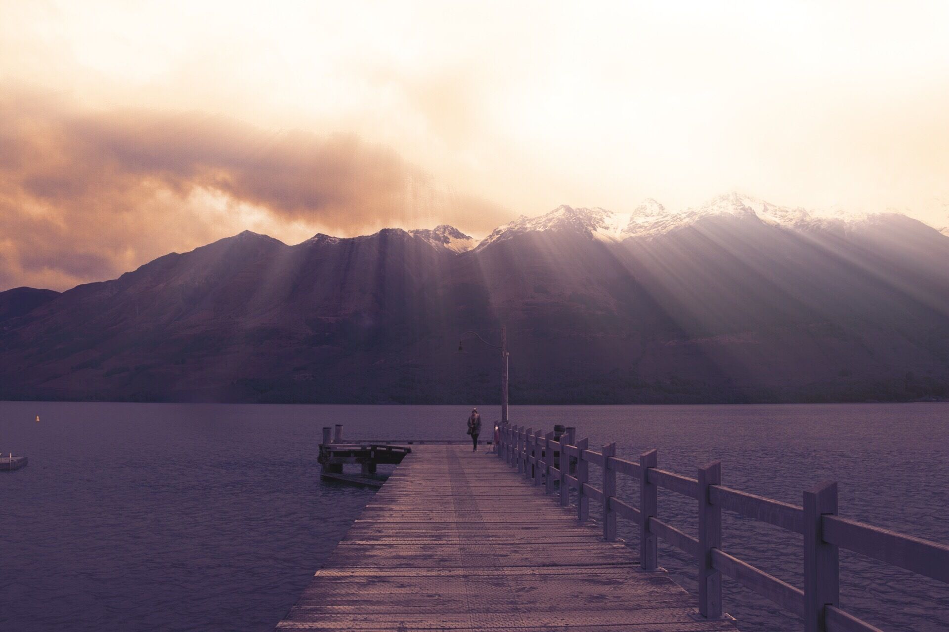 Un muelle de madera se extiende sobre un lago tranquilo con montañas al fondo y luz solar filtrándose entre las nubes.