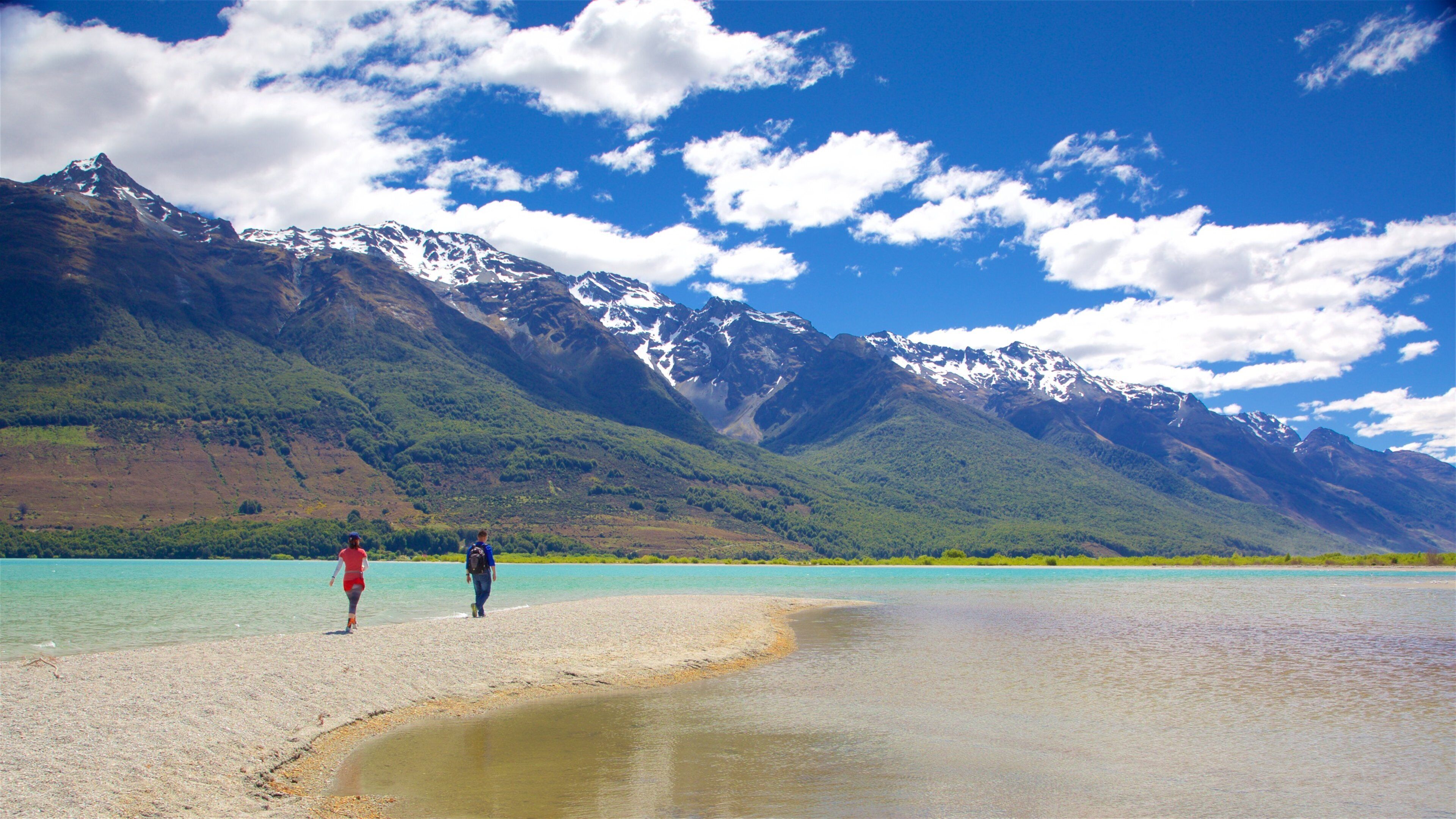 A beach with turquoise water, people walking, and snow-capped mountains in the background.